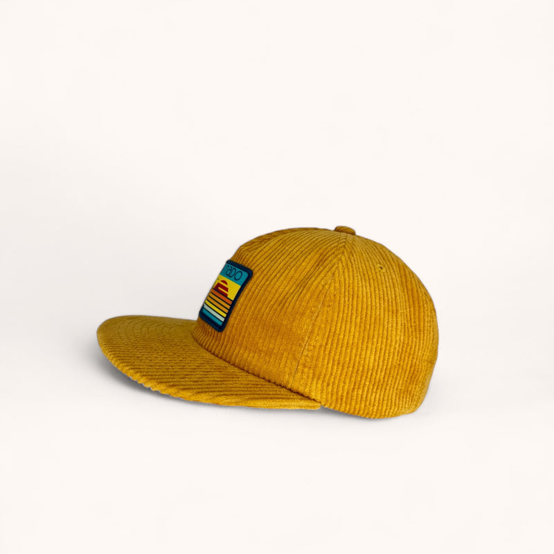 Side angle of corduroy flat bill surf hat in mustard yellow with a woven surfboard patch 