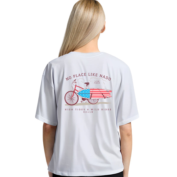 woman wearing a white, soft cotton, no place like nado tshirt with a bike carrying an american flag surfboard